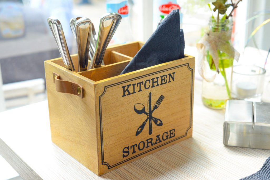 A wooden box with printed words "KITCHEN STORAGE" containing utensils and a blue napkin