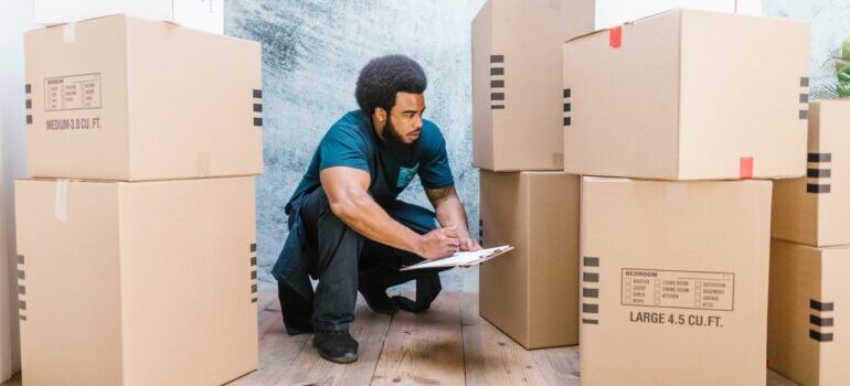 A man working as a mover in a moving companies, squatting next to bunch of cardboard boxes and writing something on the paper.