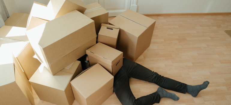 a man lying below cardboard boxes to represent one of the questions to ask before you move out of state, whether or not you should move your entire house